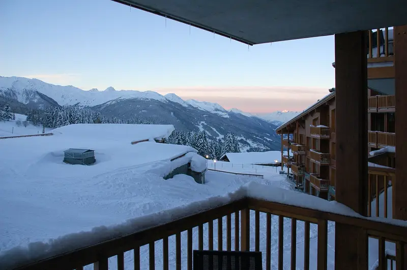 Dawn over deep snow, forest and pink-tinged peaks, seen from a wooden balcony in Arc 1800