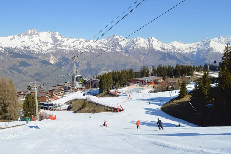 Skiers on a sunny groomed piste leading down to the Arc 1800 buildings and the Transarc gondola, with snowy Tarentaise peaks behind