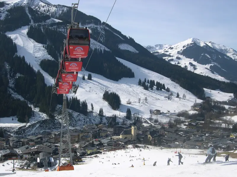 Red Kohlmais gondola cabins rising above the snowy village of Saalbach, with busy nursery slopes and ski mountains behind